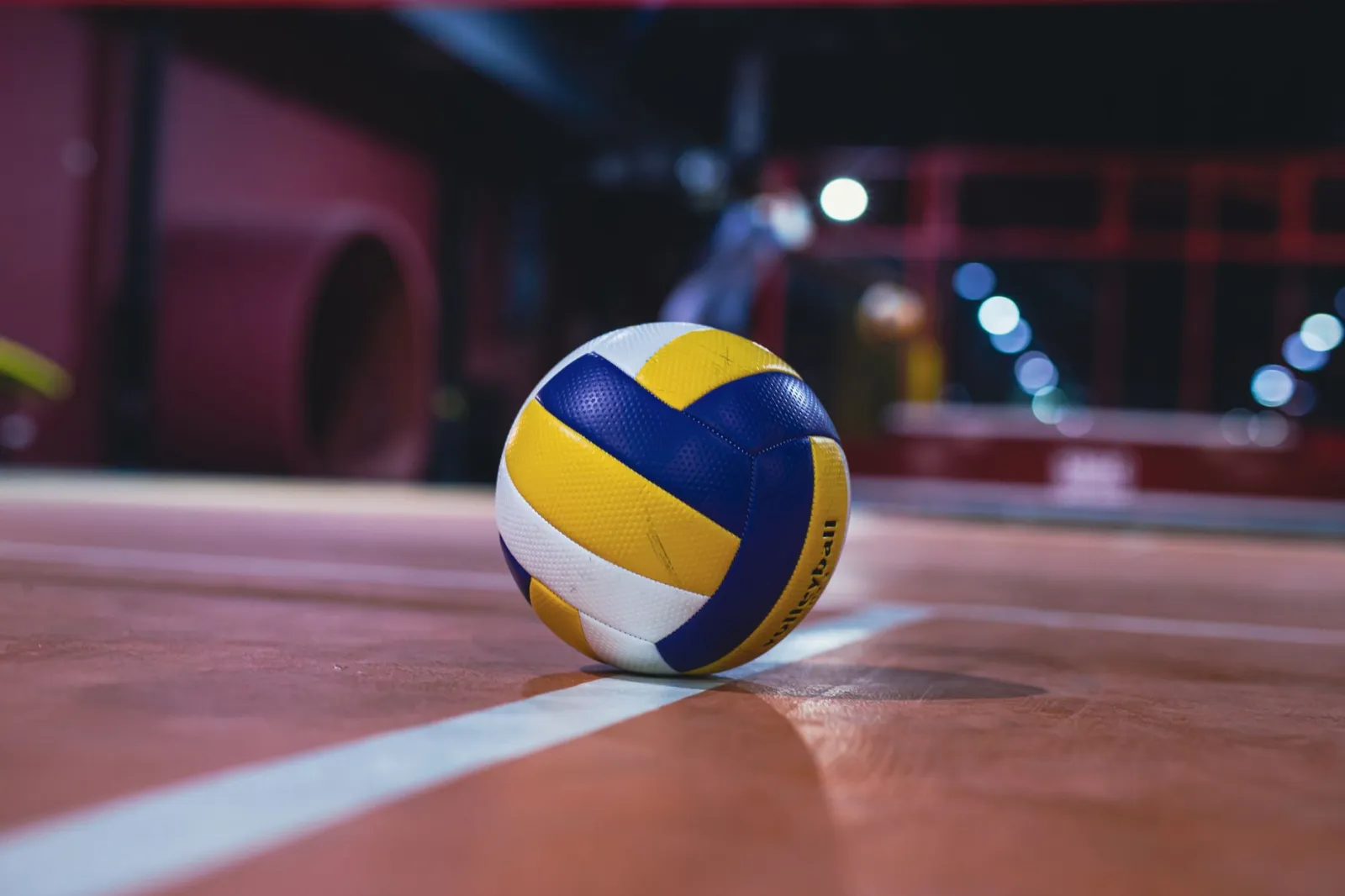 Yellow and white volleyball resting on an indoor volleyball court with bright lights.